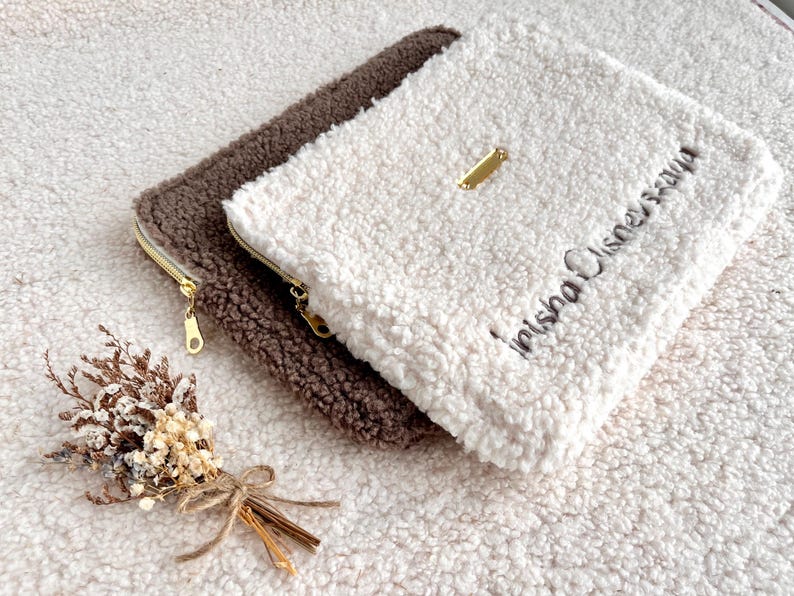 May include: Two fluffy, zippered pouches in brown and white, with gold-toned zippers and a small gold tag. The white pouch has embroidered text. A small bundle of dried flowers rests nearby.