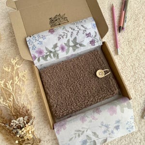 Puede incluir: Un artículo marrón texturizado con un botón de madera se encuentra en una caja de cartón forrada con papel de seda floral. La caja está abierta, revelando el contenido. Se ven un lápiz y bolígrafos.
