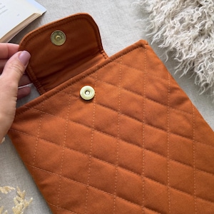 May include: A quilted rust-colored fabric sleeve featuring a diamond-stitched pattern and a gold-toned magnetic snap closure. The rectangular pouch has a flap top that secures to the main body with a metal button mechanism.