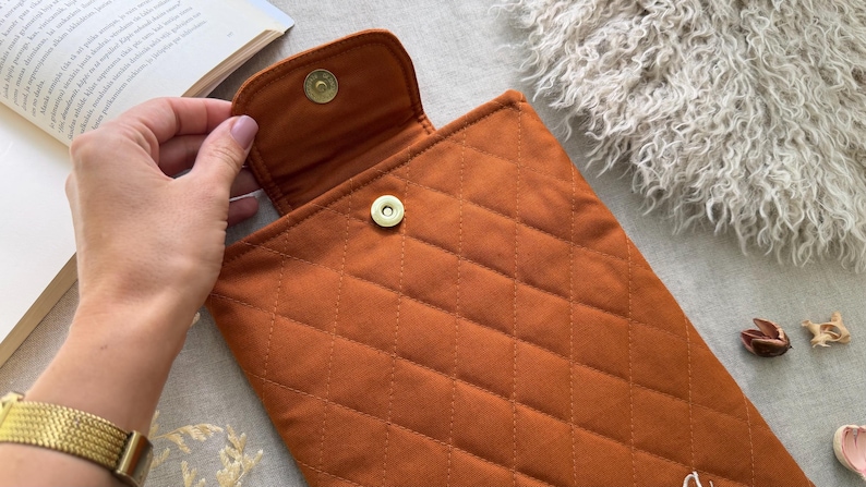 May include: A quilted rust-colored fabric sleeve featuring a diamond-stitched pattern and a gold-toned magnetic snap closure. The rectangular pouch has a flap top that secures to the main body with a metal button mechanism.
