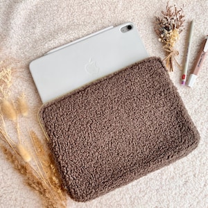 May include: A brown, textured tablet sleeve with a zipper closure, holding a white tablet. The sleeve is made of a soft, plush material. A pencil, pen, and dried flowers are next to the tablet. The items are on a white, fluffy surface.