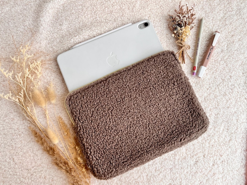 May include: A brown, textured tablet sleeve with a zipper closure, holding a white tablet. The sleeve is made of a soft, plush material. A pencil, pen, and dried flowers are next to the tablet. The items are on a white, fluffy surface.
