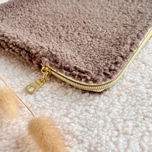 May include: A rectangular, brown, fluffy pouch with a gold zipper and pull. The pouch is made of a soft, textured material. The zipper is gold-colored. The pouch is lying on a white, fluffy surface.