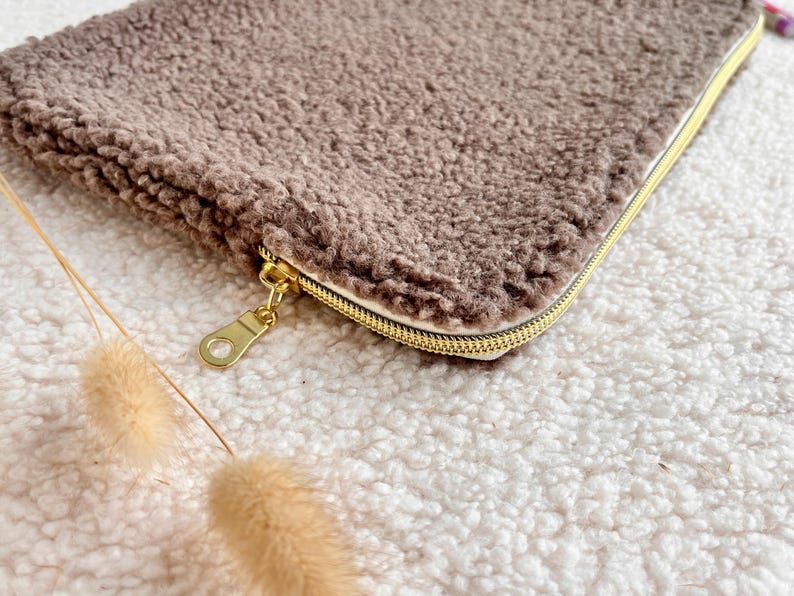 May include: A rectangular, brown, fluffy pouch with a gold zipper and pull. The pouch is made of a soft, textured material. The zipper is gold-colored. The pouch is lying on a white, fluffy surface.