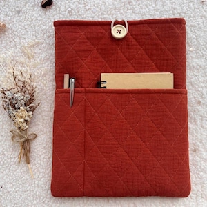 May include: A rectangular, quilted, rust-coloured fabric sleeve featuring a diamond-stitched pattern. The sleeve has a front pocket containing a small spiral-bound notebook and a silver pen, secured at the top with a single beige button and loop.