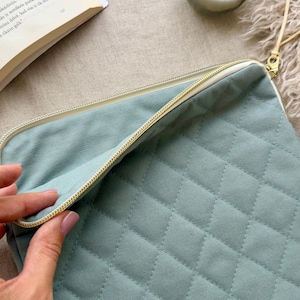 May include: A sage green quilted laptop sleeve featuring a diamond-stitched pattern, a cream-colored zipper closure, and a matching leather-like zipper pull. The rectangular case is partially open, revealing a plain interior lining.