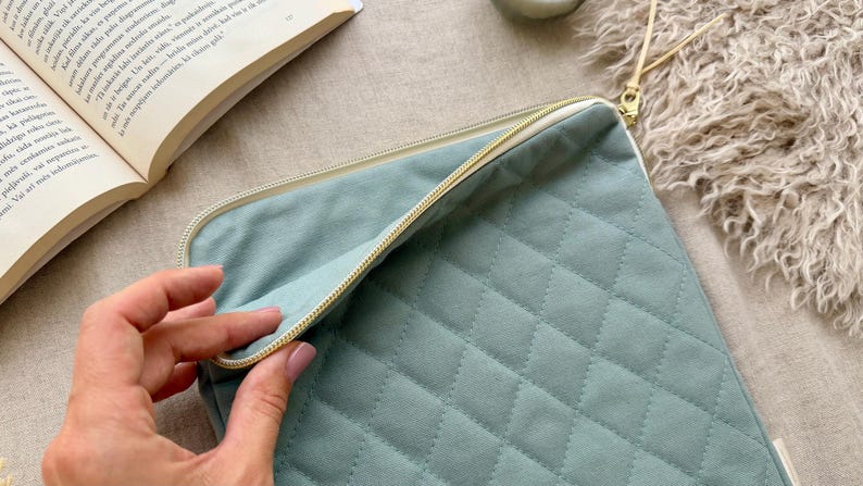 May include: A sage green quilted laptop sleeve featuring a diamond-stitched pattern, a cream-colored zipper closure, and a matching leather-like zipper pull. The rectangular case is partially open, revealing a plain interior lining.
