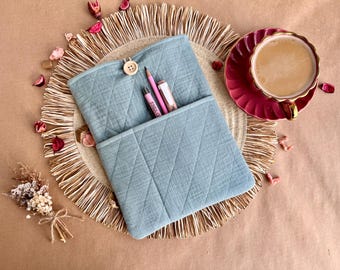 May include: A light blue quilted fabric pencil case with a wooden button closure. The case contains pink pencils and a pen. A cup of coffee in a red teacup sits nearby on a woven placemat, with dried flowers and petals scattered around.