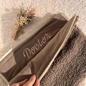 May include: A brown zippered case with the word "Doctor" embroidered on the inside. The case is open, revealing a soft, textured interior. A small bouquet of dried flowers rests nearby. The case is on a fluffy, light-colored surface.