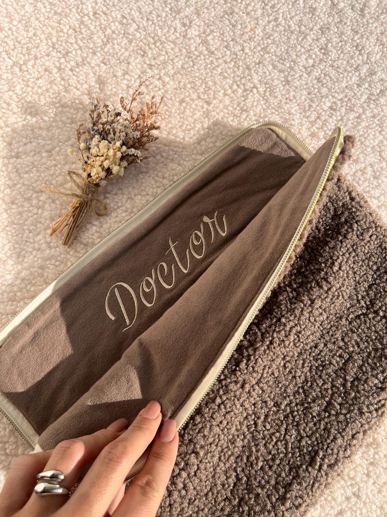 May include: A brown zippered case with the word "Doctor" embroidered on the inside. The case is open, revealing a soft, textured interior. A small bouquet of dried flowers rests nearby. The case is on a fluffy, light-colored surface.