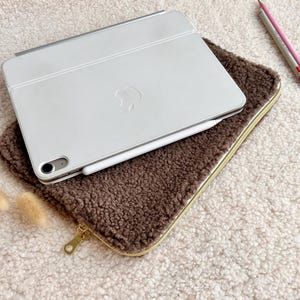 May include: A white tablet with a silver keyboard cover and a white stylus rests on a brown, fuzzy case with a gold zipper. Two pencils, one pink and one gray, are next to the case.