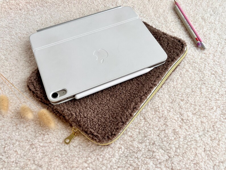 May include: A white tablet with a silver keyboard cover and a white stylus rests on a brown, fuzzy case with a gold zipper. Two pencils, one pink and one gray, are next to the case.
