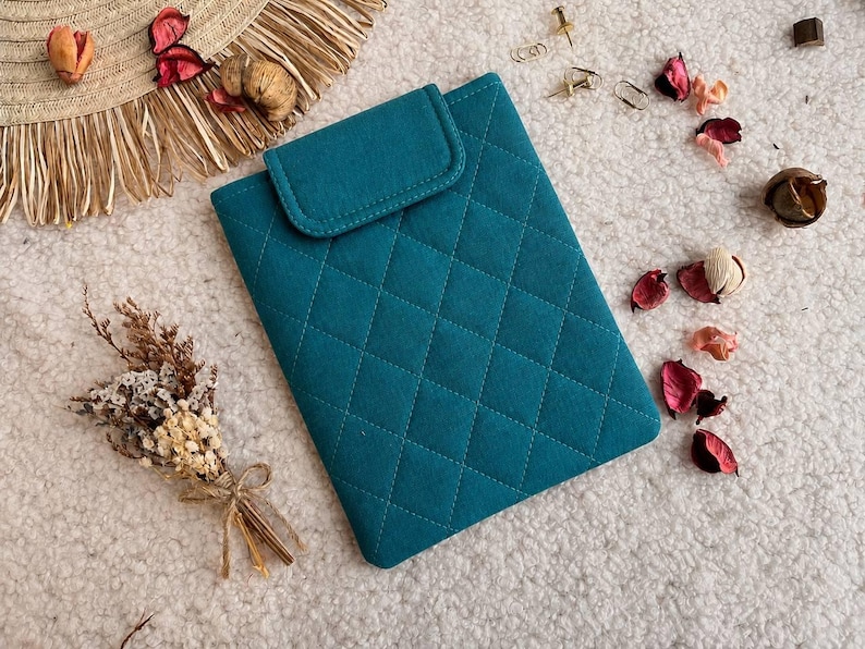 May include: A rectangular teal tablet sleeve featuring a quilted diamond pattern and a fold-over flap closure. The sleeve is constructed from a soft fabric material and displays a consistent stitched grid design across its entire front surface.