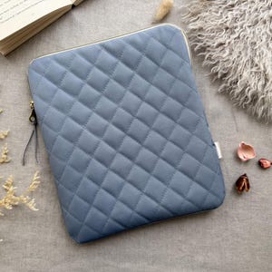 May include: A rectangular, slate blue laptop sleeve featuring a quilted diamond pattern across the front surface. The sleeve includes a gold-toned metal zipper closure with a thin, dark gray leather pull tab attached to the zipper slider.