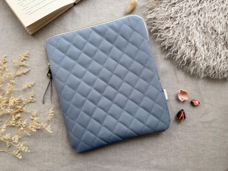 May include: A rectangular, slate blue laptop sleeve featuring a quilted diamond pattern across the front surface. The sleeve includes a gold-toned metal zipper closure with a thin, dark gray leather pull tab attached to the zipper slider.