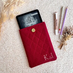 Könnte beinhalten: Eine rote, gesteppte E-Reader-Hülle mit goldenem Druckknopfverschluss, die einen E-Reader mit dem Buch "The Girl in Cabin 13" enthält. Die Hülle hat die gestickten Initialen "K.J.". Ein Stift, ein Bleistift und Trockenblumen sind ebenfalls vorhanden.