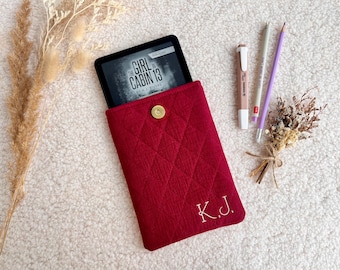 May include: A crimson quilted e-reader case with a gold press stud, holding an e-reader displaying the book "The Girl in Cabin 13." The case has the embroidered initials "K.J." A pen, pencil, and dried flowers are also present.