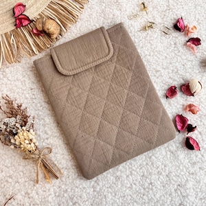 May include: A tan quilted tablet case with a flap closure. The case has a diamond pattern and is surrounded by decorative items, including a straw hat, dried flowers, and petals. The case is designed to protect a tablet.