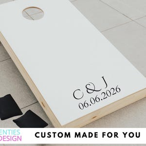 May include: A white cornhole board with a circular hole at the top and natural wood sides. The board features black text reading C & J and the date 06.06.2026. Two solid black bean bags are positioned on the ground next to the wooden game board.