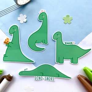 May include: Four green dinosaur stickers with different expressions. One dinosaur is eating a slice of pizza, another is sleeping, another is saying "RAWR" and the last one has hearts above its head.