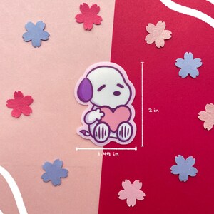 May include: A sticker featuring a cartoon dog with a purple ear and a pink heart. The sticker is 1.49 inches wide and 2 inches tall. The background is split between pink and red, with scattered flower-shaped cutouts in pink and blue.