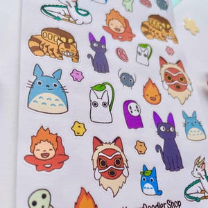 May include: A sheet of stickers featuring characters from Studio Ghibli films. The stickers are colorful and feature characters like Totoro, No Face, and Kiki's Delivery Service. The sheet is titled "Ghibli Friends" and includes the shop name "The Happy Doodler Shop".