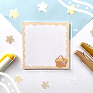 May include: A set of 3x3 inch beige sticky notes with a scalloped edge and a pink heart pattern. The notes feature a cartoon illustration of a brown wicker basket filled with bread.