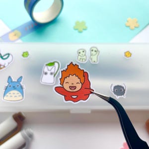May include: A clear plastic case with a variety of colorful cartoon stickers, including a red character with orange hair, a white cat, a blue creature with large ears, and a gray creature with a single eye. The stickers are being placed in the case with tweezers.
