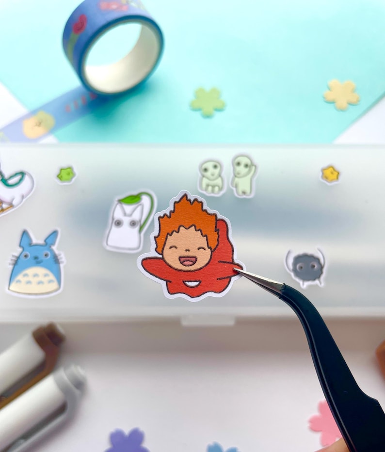 May include: A clear plastic case with a variety of colorful cartoon stickers, including a red character with orange hair, a white cat, a blue creature with large ears, and a gray creature with a single eye. The stickers are being placed in the case with tweezers.