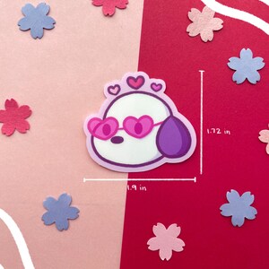 May include: A sticker featuring a cartoon dog with heart-shaped sunglasses and a crown of hearts. The sticker is approximately 1.9 inches wide and 1.72 inches tall. Scattered around the sticker are pink and blue flower cutouts against a pink and red background.