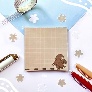 May include: A brown gridded sticky note with a cartoon illustration of a brown Bigfoot walking across a path. The note is 3 inches wide and 3 inches tall. There are small white flower stickers on a white background.