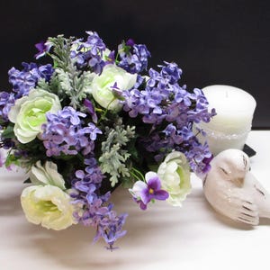 May include: A floral arrangement featuring purple, white, and green flowers. A white candle and a white ceramic bird are also present. The flowers include lavender, roses, and other blooms, creating a soft, natural aesthetic.