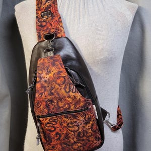 May include: Black leather sling bag with a patterned strap and front pocket. The pattern features orange, red, and black swirling designs. The bag has a zippered pocket and a silver-tone metal clasp.
