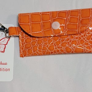 Könnte beinhalten: Orangefarbene Kunstleder-Geldbörse mit Druckknopfverschluss und silberfarbenem Verschluss. Ein weißes Etikett mit roter Schmetterlingsgrafik und dem Text "love, joshua limited edition" ist angebracht. Ein weißes Band mit Schmetterlingsgrafik ist ebenfalls angebracht.