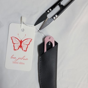 May include: Black and silver thread snips with a pink thread cutter in a black leather sheath. A white tag with a red butterfly graphic and the text "love, joshua limited edition" is attached.