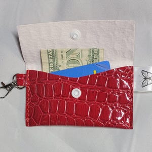 Könnte beinhalten: Eine rote, strukturierte Kunstleder-Geldbörse mit silbernem Verschluss und weißem Druckknopfverschluss. Die Geldbörse ist geöffnet und zeigt einen Zwanzig-Dollar-Schein und eine blaue Karte. Ein weißes Band mit einer Schmetterlingsillustration ist befestigt.