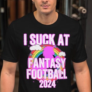 Puede incluir: Camiseta negra con un gráfico de unicornio rosa y arcoíris. El texto "I suck at Fantasy Football 2024" está impreso en la camiseta.