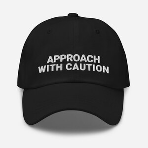 Puede incluir: Una gorra de béisbol negra con texto bordado en blanco que dice "APPROACH WITH CAUTION".
