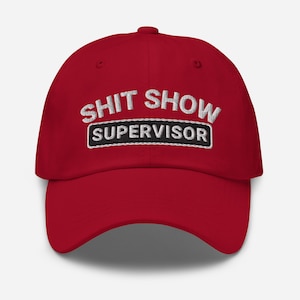 以下が含まれることがあります： 白い刺繍で「Shit Show Supervisor」と書かれた赤いベースボールキャップ。