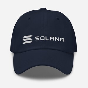 May include: Navy blue baseball cap with the word "SOLANA" embroidered in white. The cap features the Solana logo on the left side. A classic, adjustable hat for everyday wear.