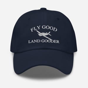 Könnte beinhalten: Marineblaue Baseballkappe mit weiß gesticktem Text "FLY GOOD LAND GOODER" und einer kleinen Flugzeuggrafik.