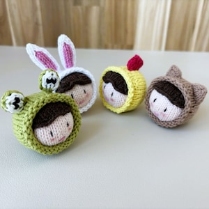 Può includere: Quattro bambole di lana con diversi cappelli a tema animale. Le bambole indossano un cappello da coniglio, un cappello da rana, un cappello da fiore e un cappello da gatto. Le bambole hanno i capelli castani e le guance rosa.