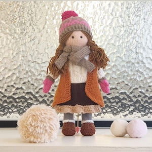Op de afbeelding: Een gebreide pop die een bruine hoed met een roze pompon, een bruine sjaal, een oranje vest, een bruine rok en bruine laarzen draagt. De pop houdt een sneeuwbal in elke hand. De pop staat op een wit oppervlak voor een raam.