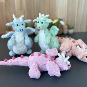 Puede incluir: Tres juguetes de dragón de punto. Uno es azul claro, otro es verde claro y otro es rosa. Todos están sentados sobre una superficie gris.