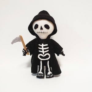 Op de afbeelding: Een gebreide Grim Reaper-pop met een zwarte capuchonmantel en een kleine zeis. De pop heeft een wit schedelgezicht met zwarte oogkassen en een zwarte capuchon. Het skelet is wit gedetailleerd op de zwarte mantel.