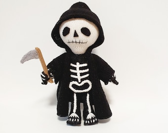 May include: A knitted Grim Reaper doll, featuring a black hooded cloak and a small scythe. The doll has a white skull face with black eye sockets and a black hood. The skeleton is detailed in white on the black cloak.