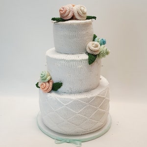 Könnte beinhalten: Eine dreistöckige gestrickte Hochzeitstorte, verziert mit gehäkelten Blumen in Rosa-, Grün- und Blautönen.