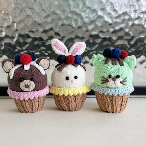 May include: Three crocheted stuffed animals shaped like cupcakes. The first is a brown bear with pink frosting, the second is a white bunny with yellow frosting, and the third is a green cat with blue frosting.