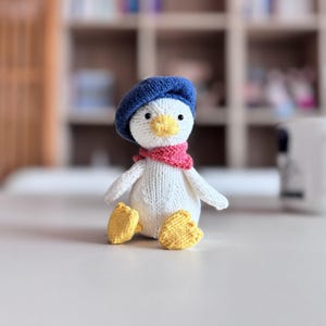May include: A knitted duckling toy, handmade with white yarn. It has a yellow beak and feet, a blue beret, and a pink scarf. The toy is sitting on a white surface.