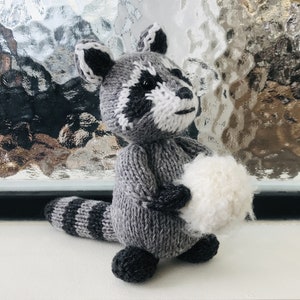May include: A gray and white knitted raccoon holding a white fluffy ball. The raccoon is standing on a white surface in front of a window.
